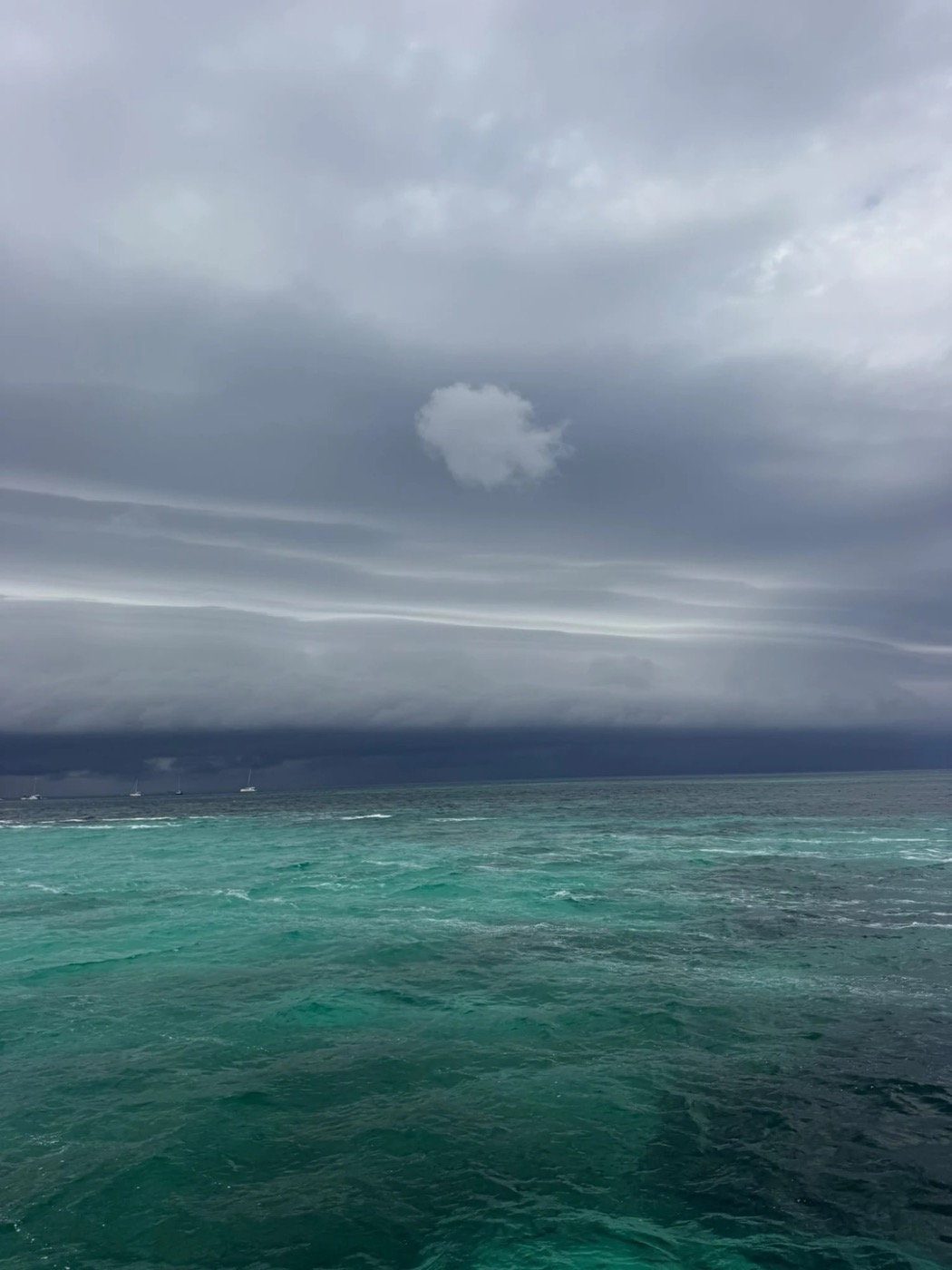 Een buienlijn boven een ankerplaats: grijze wolkenbanden boven turquoise water, een paar ankerende boten aan de horizon.